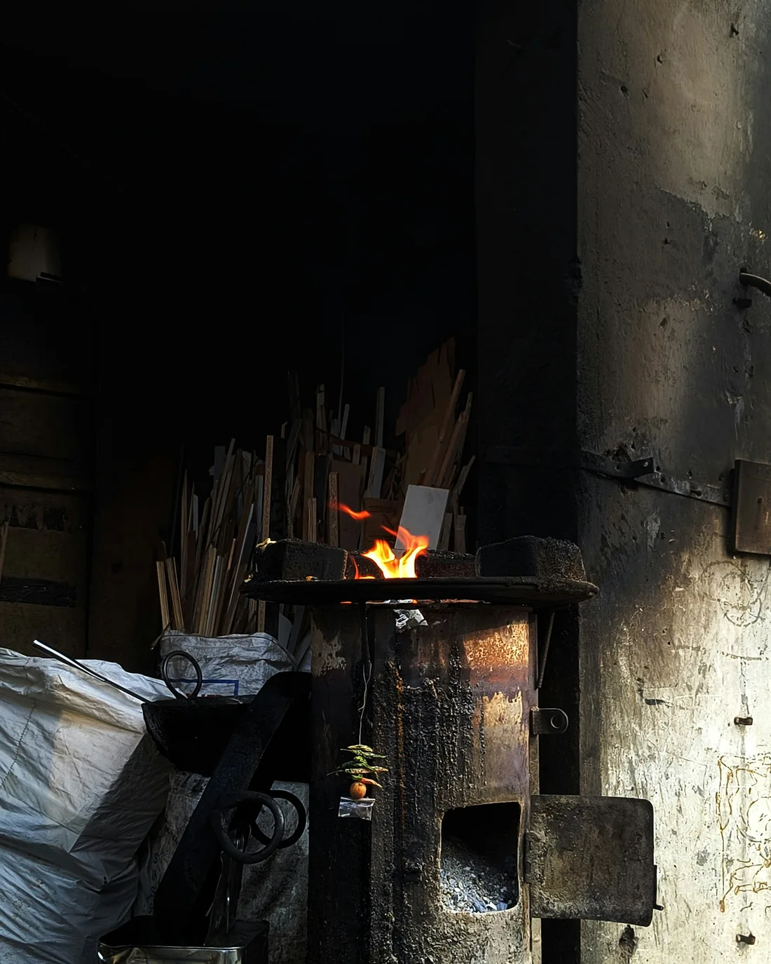 Horno de taller con la boca abierta y llama viva entre restos de leña, en un espacio de cerámica en penumbra